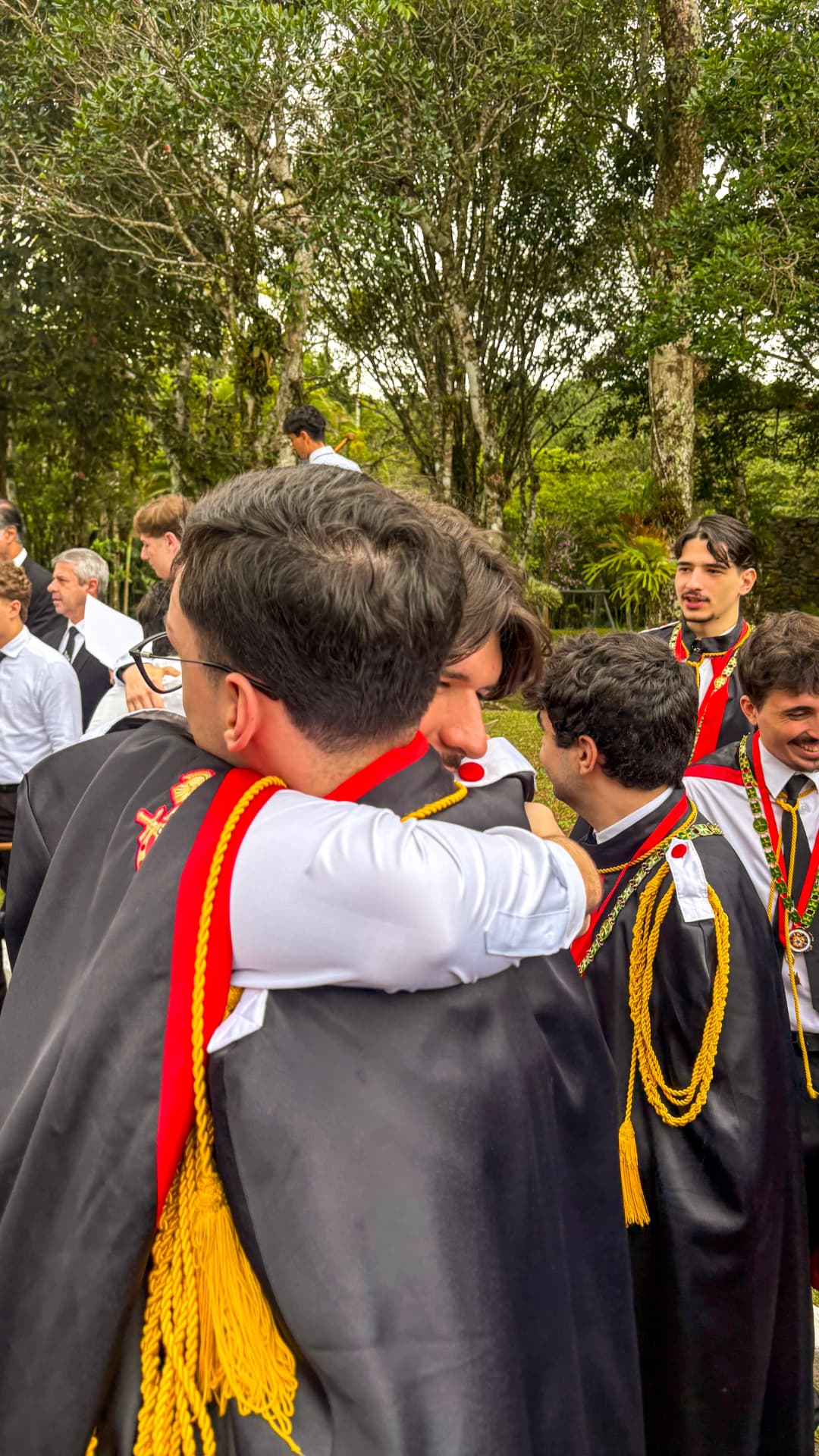 DeMolays abraçados em reunião ao ar livre, com paramentos cerimoniais pretos, vermelhos e dourados